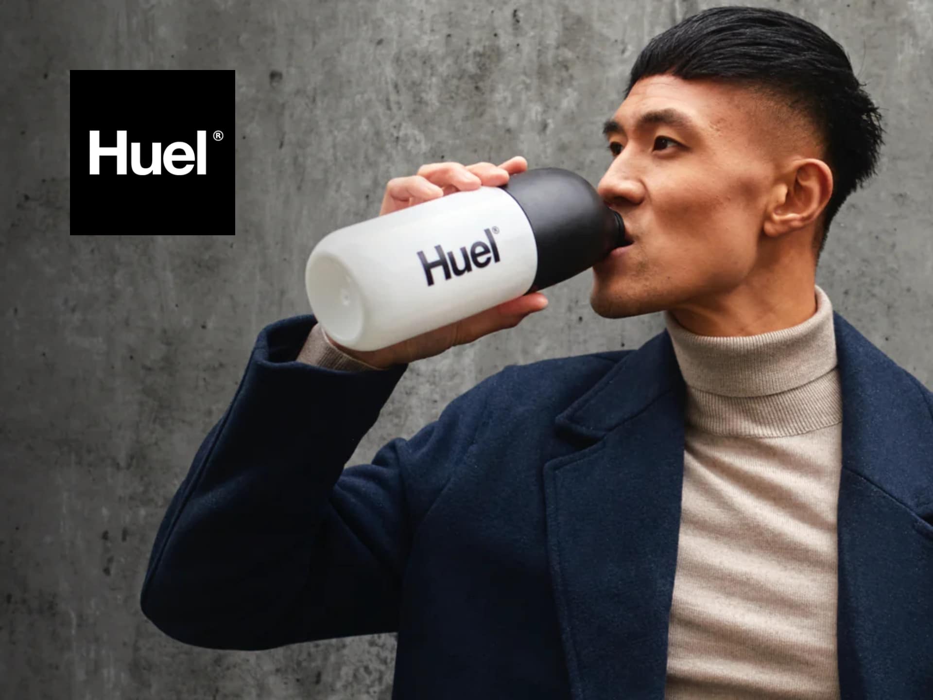 Man drinking from a black Huel shaker bottle against a concrete wall