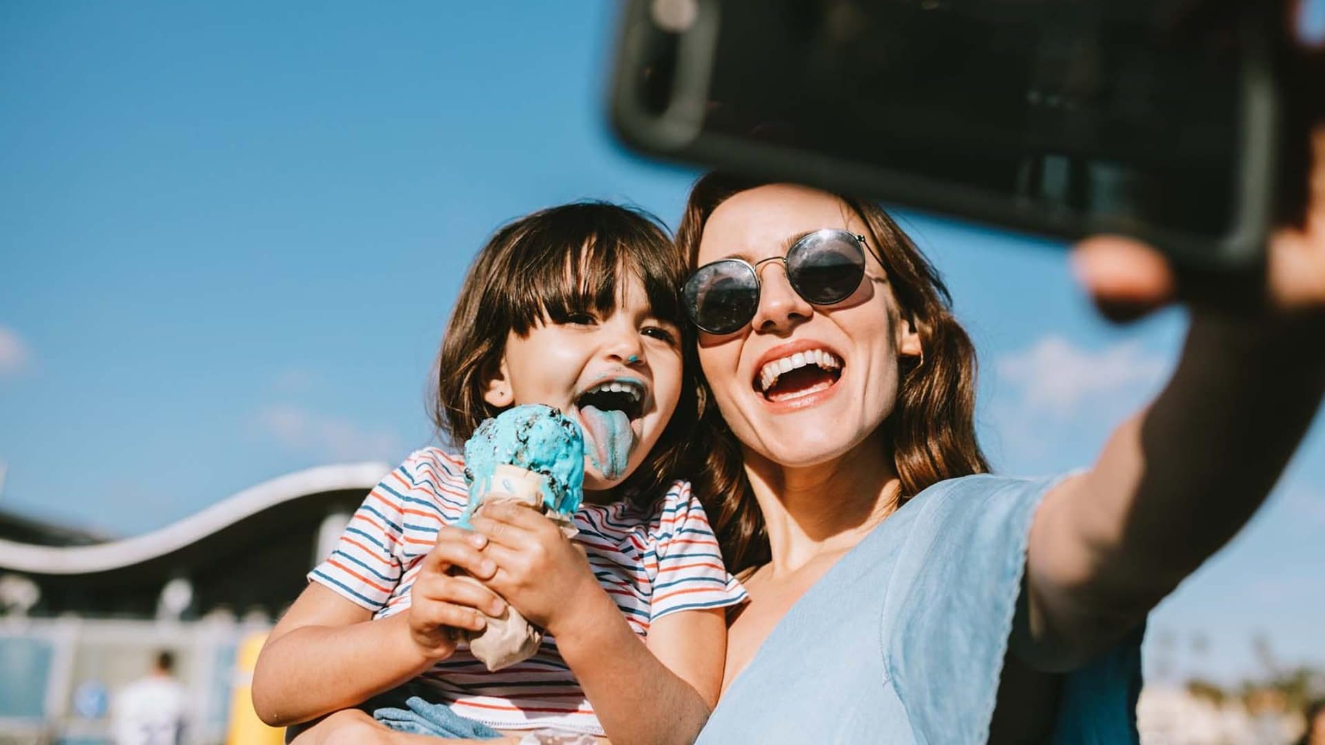 Mother and child taking a selfie while eating blue ice cream outdoors on a sunny day.