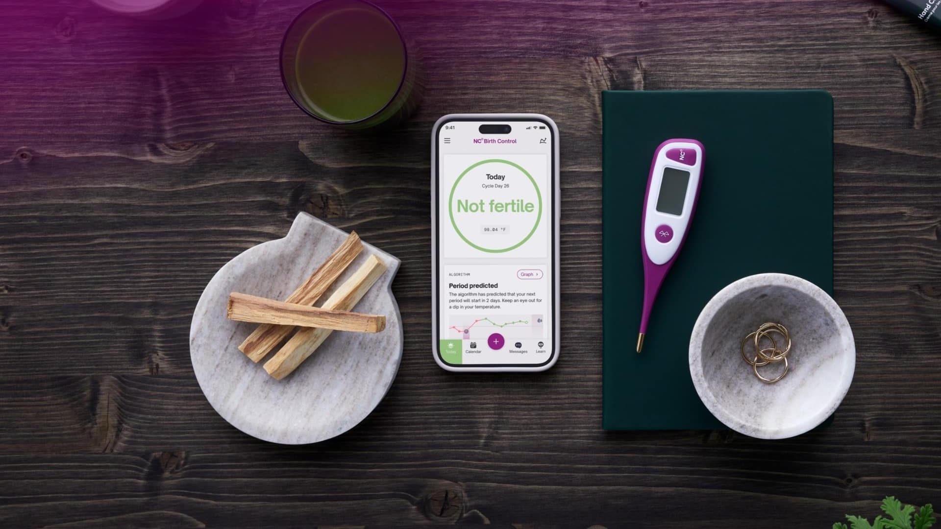 Natural Cycles thermometer and smartphone app interface on a bedside table for hormone-free contraception tracking.