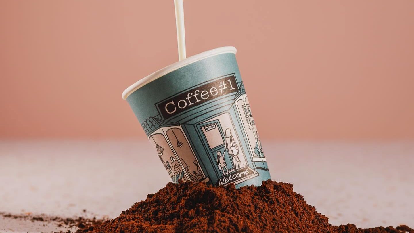A Coffee#1 branded takeaway cup resting in a pile of fresh ground coffee beans against a soft pink background.