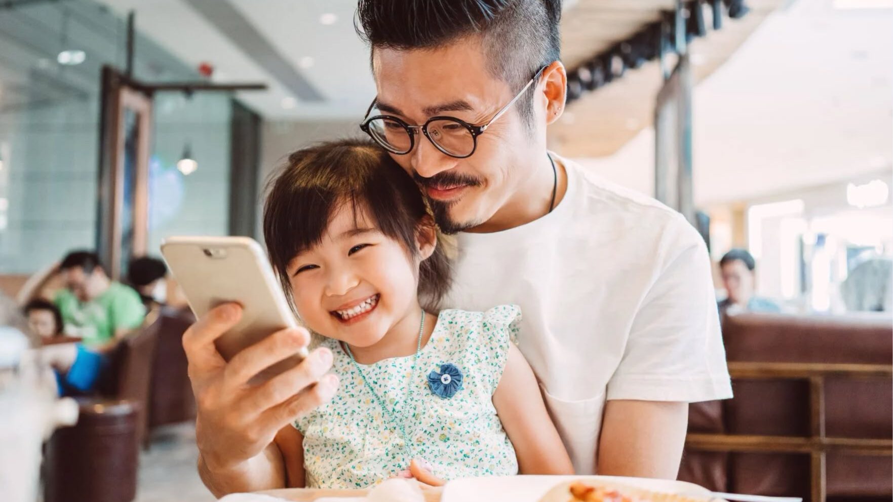 A father and daughter smiling while looking at a smartphone screen together in a bright cafe setting.