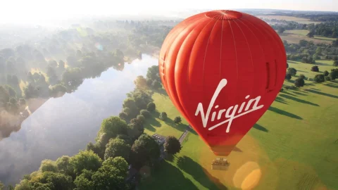 A vibrant red Virgin hot air balloon flying over the scenic British countryside during a sunrise flight experience.