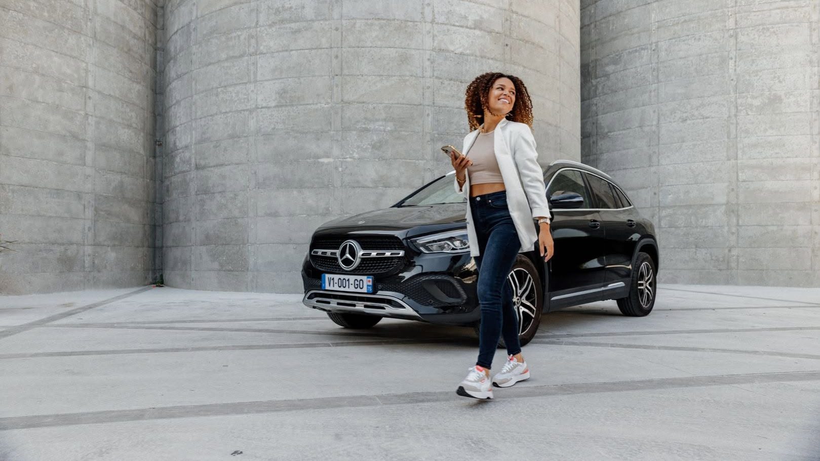Woman using smartphone to unlock a premium Mercedes rental car from Virtuo in a modern urban setting.