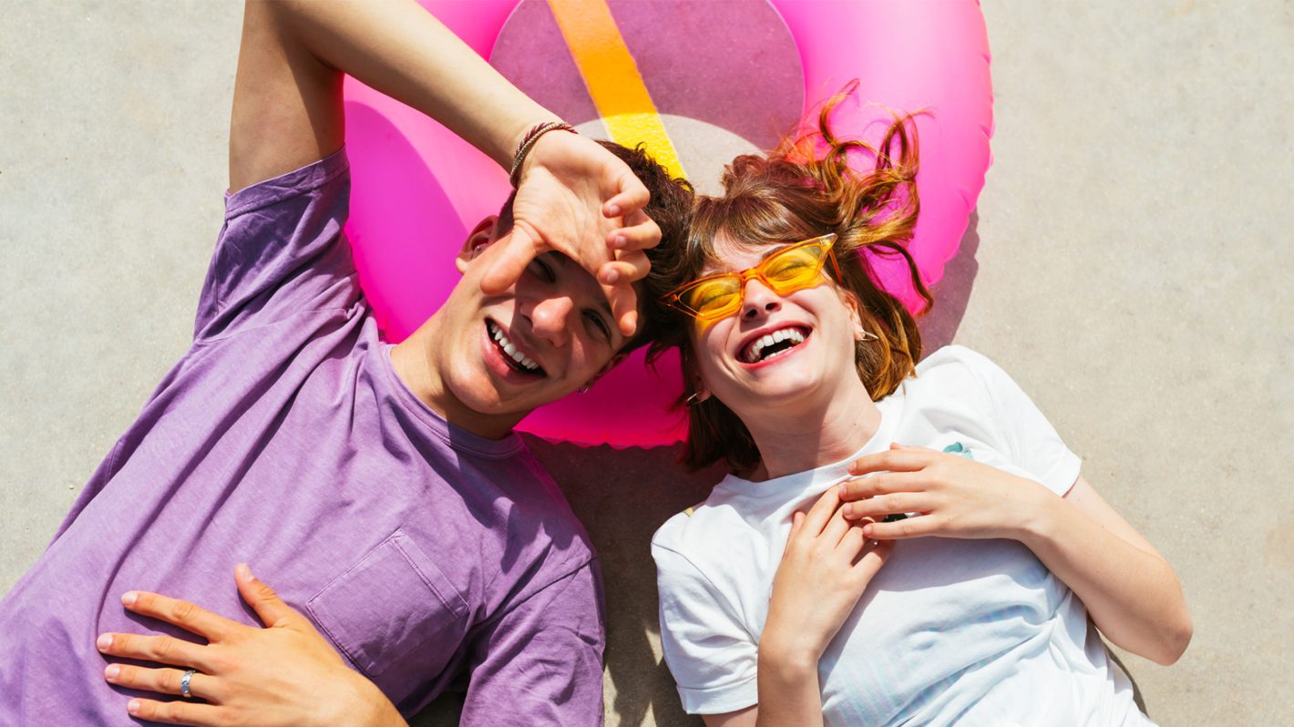Young couple laughing while lying on the ground with a bright pink inflatable ring in a sunny outdoor setting.