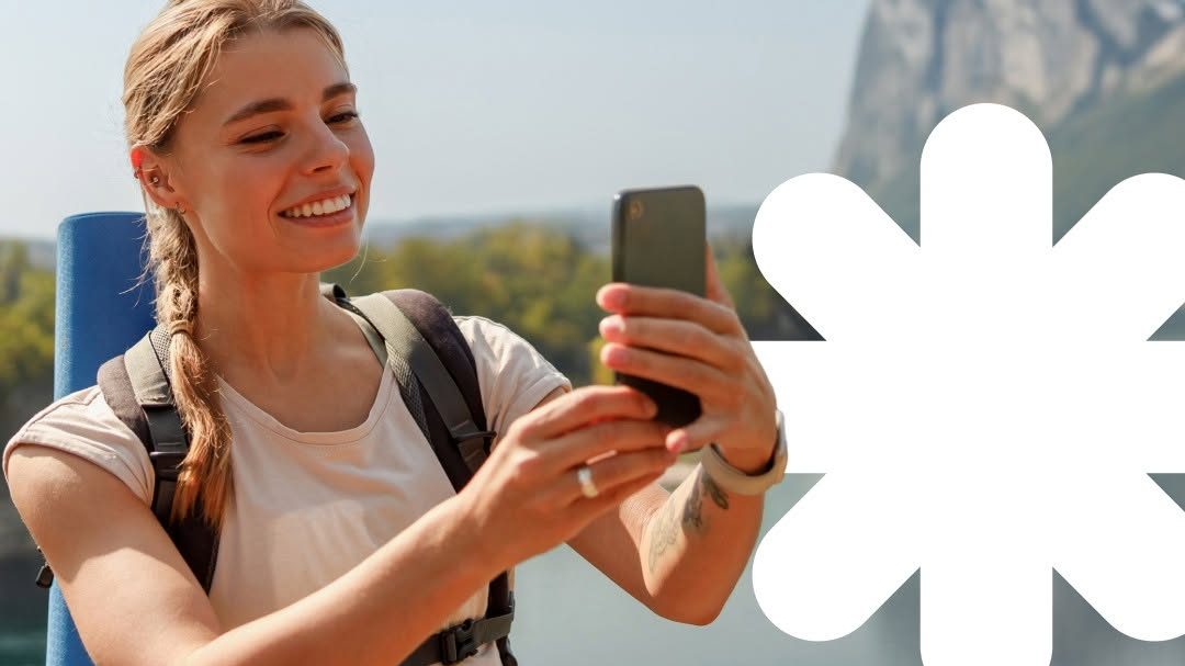 A smiling female hiker using a smartphone to capture a scenic mountain view while travelling abroad.