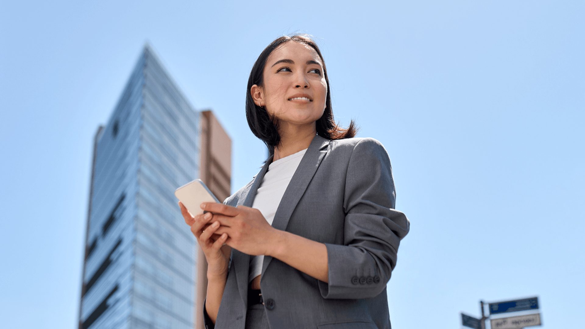 Professional woman in a grey suit holding a smartphone against a modern city skyline and clear blue sky.