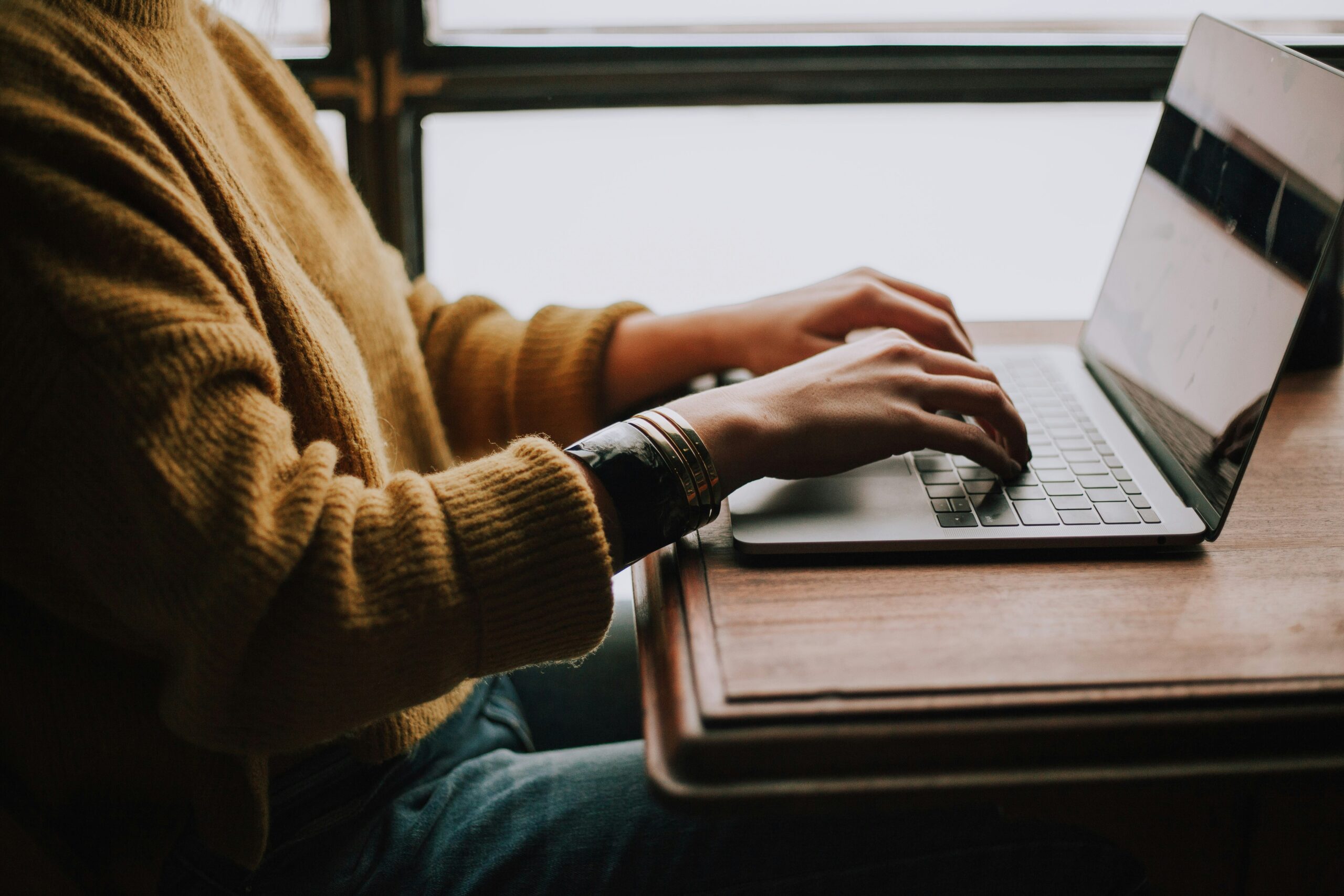 Student typing on a laptop at a wooden desk by a window
