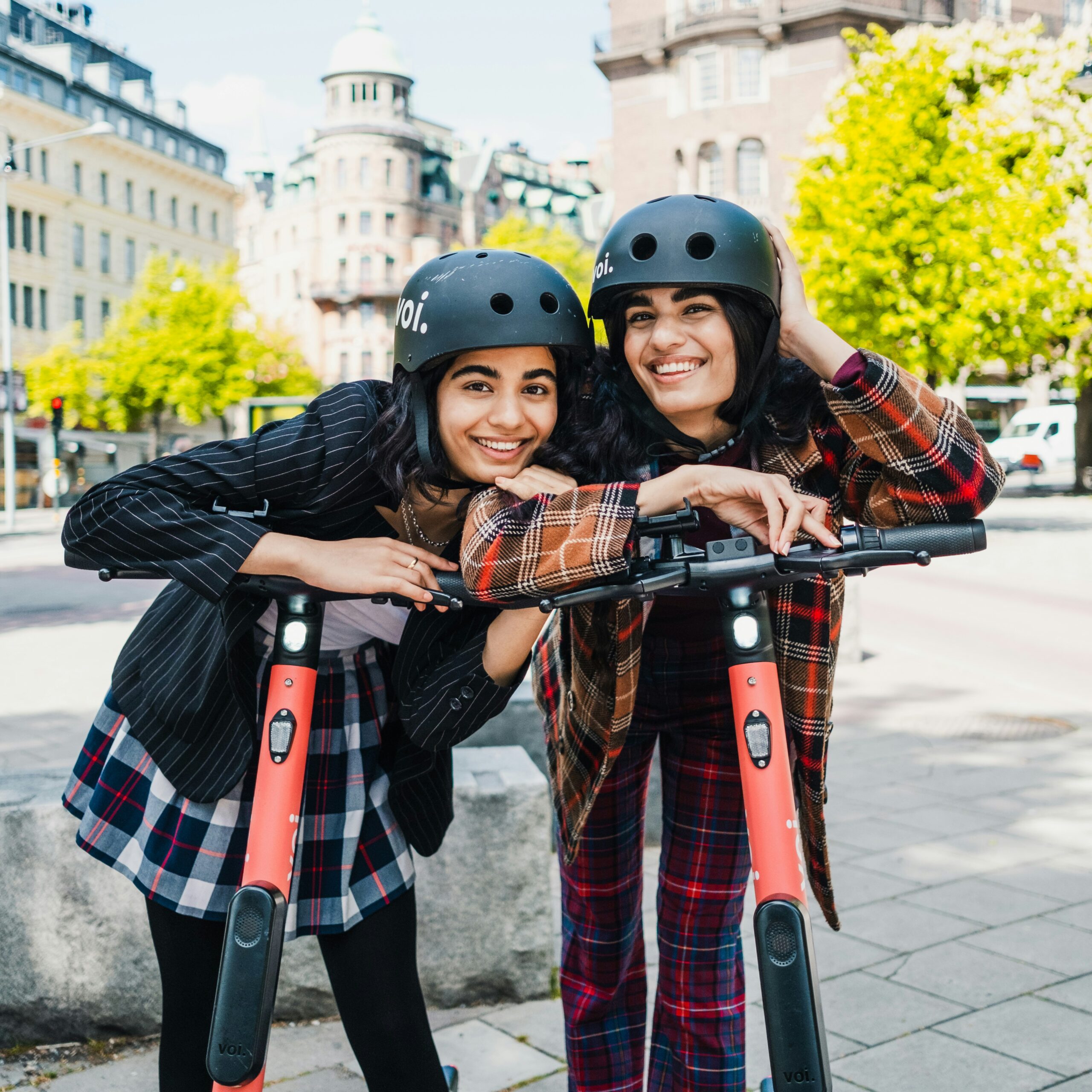 Two riders wearing helmets with coral Voi electric rental scooters on city pavement